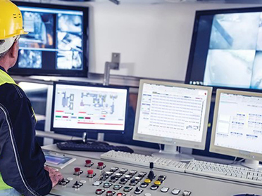 A worker in a hard hat and safety vest attentively monitors the screens.